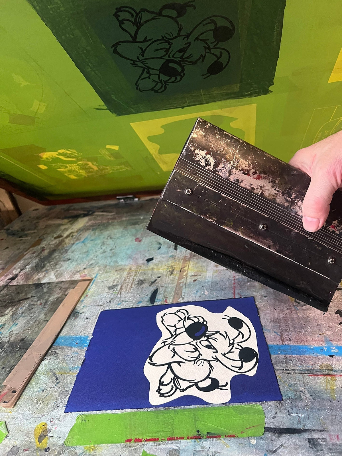 A screenprint of Dogmatix lies on the printing bed, just after the black ink has been applied. The background and his nose are blue. A squeegee is also in frame