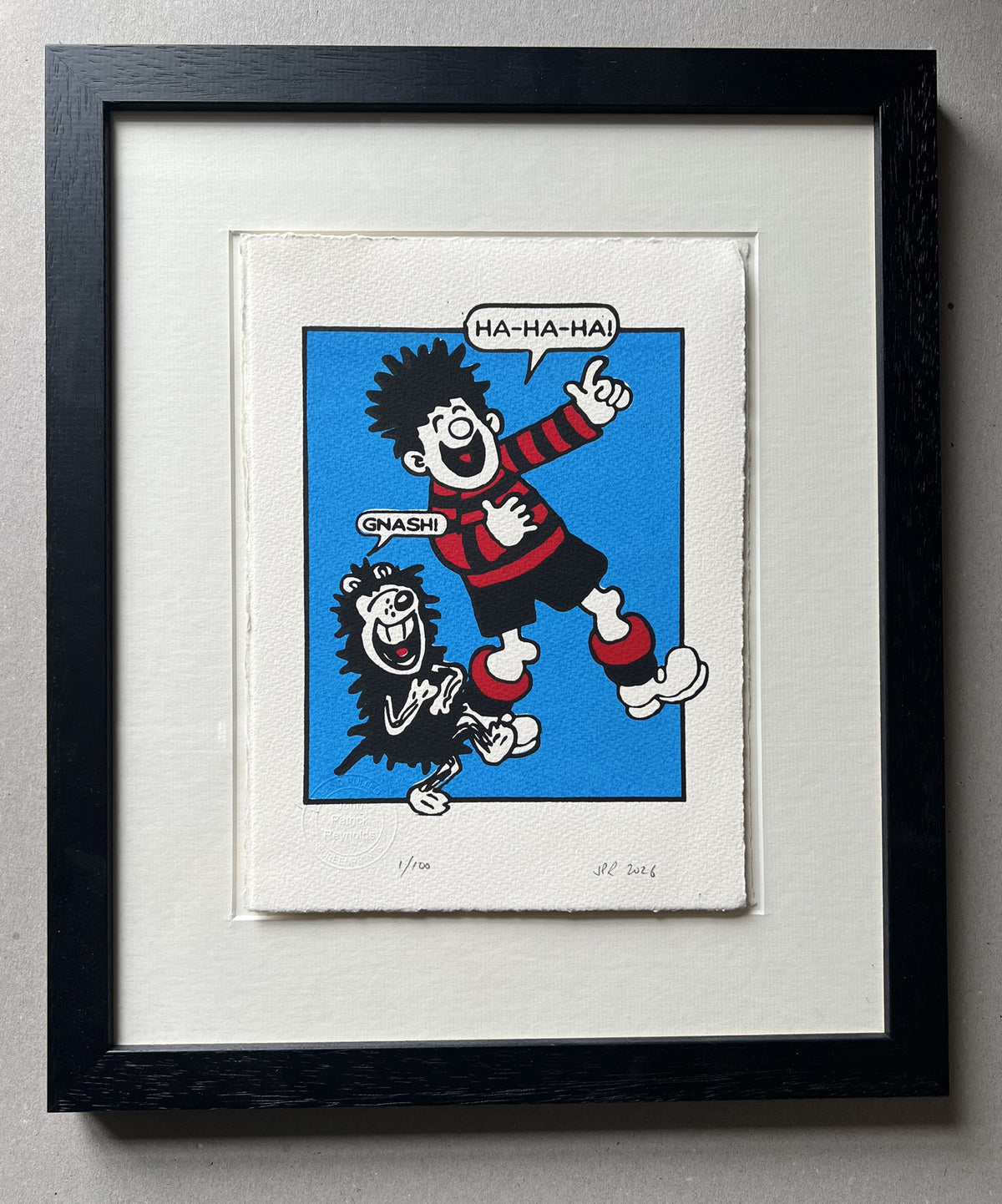 Dennis the menace from the beano and his dog gnasher are laughing at something and hold their stomachs in hilarity; he is dressed in a red and black striped football kit. In a black frame